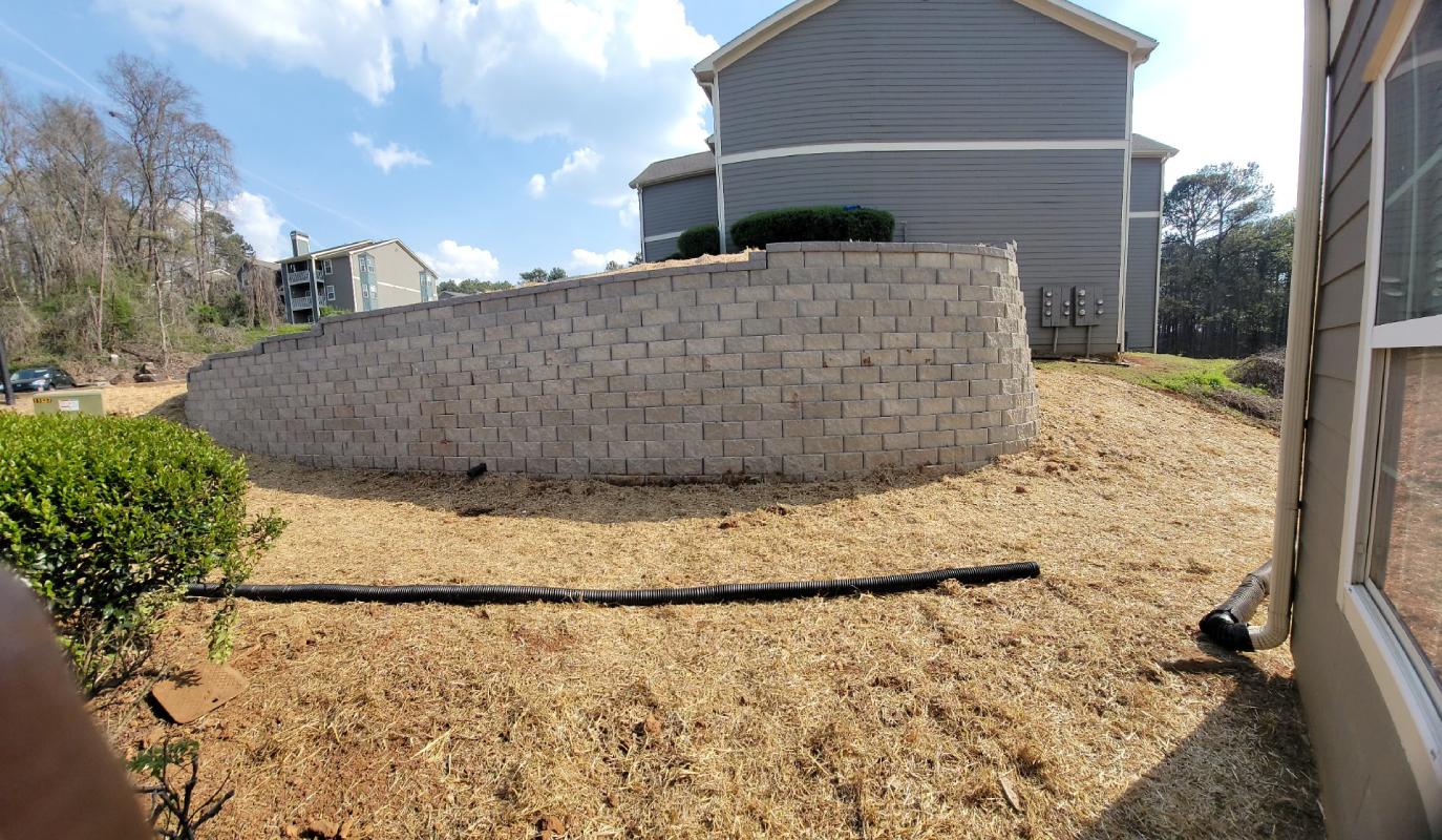 A gray brick retaining wall with a slope of straw-covered ground is in front of a two-story gray house. Leafless trees are in the background under a blue sky.